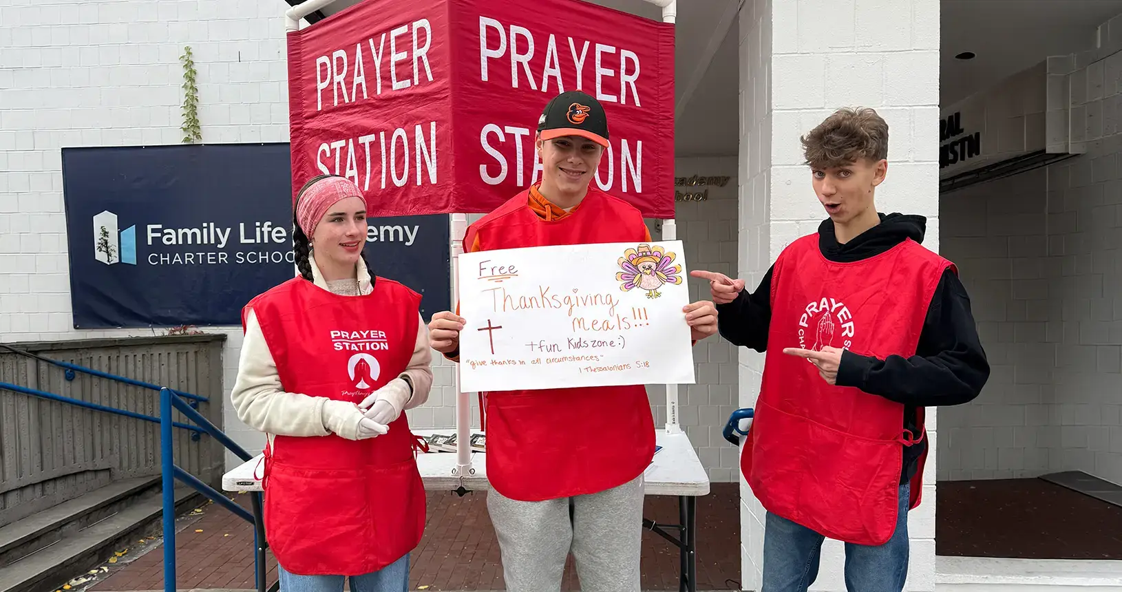Three young volunteers in red vests at a prayer station holding a sign for free Thanksgiving meals